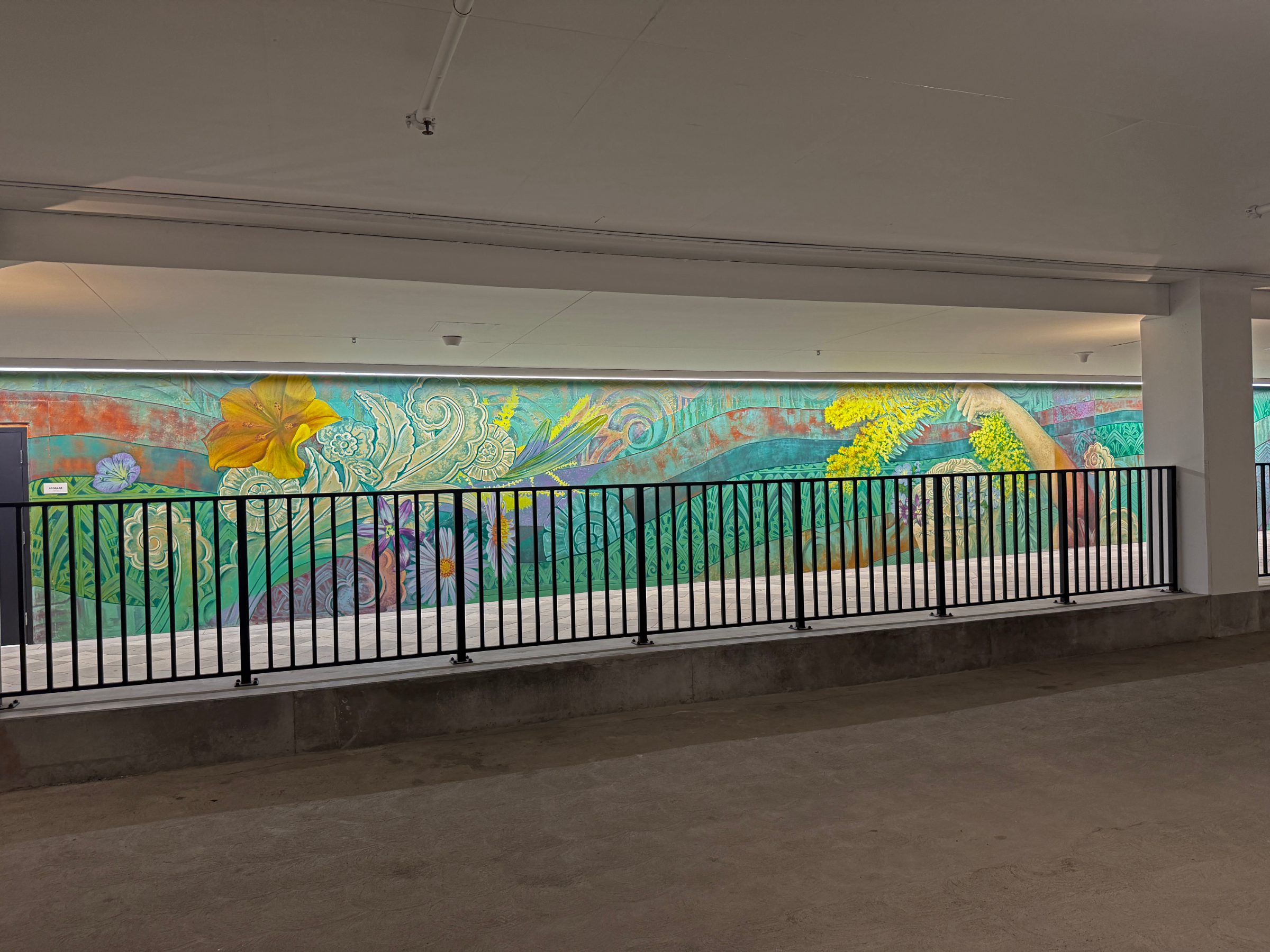 "Flourishing Structures" mural at Bloom on Third seen across the parking level beyond a black railing